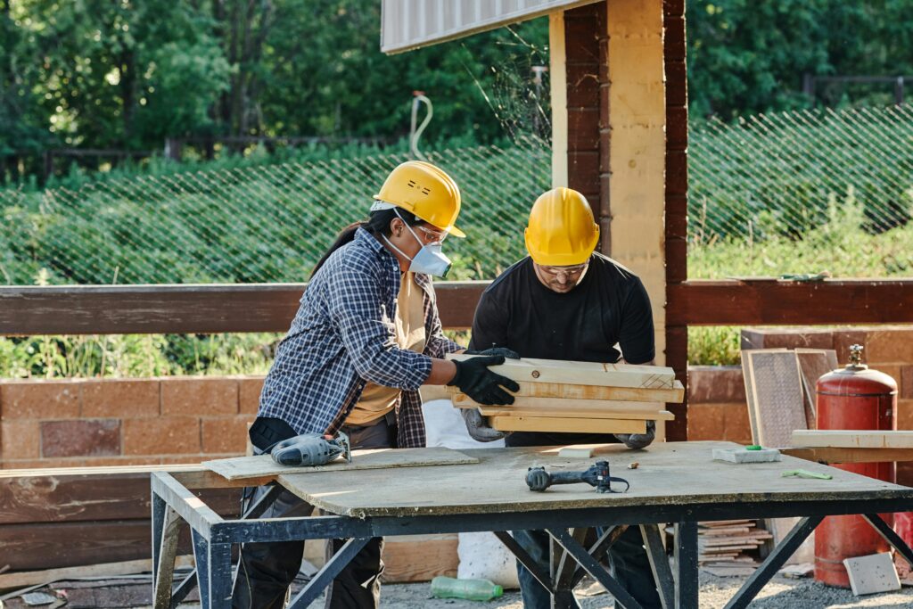Two construction workers organize wooden planks on a table at a sunny outdoor site.